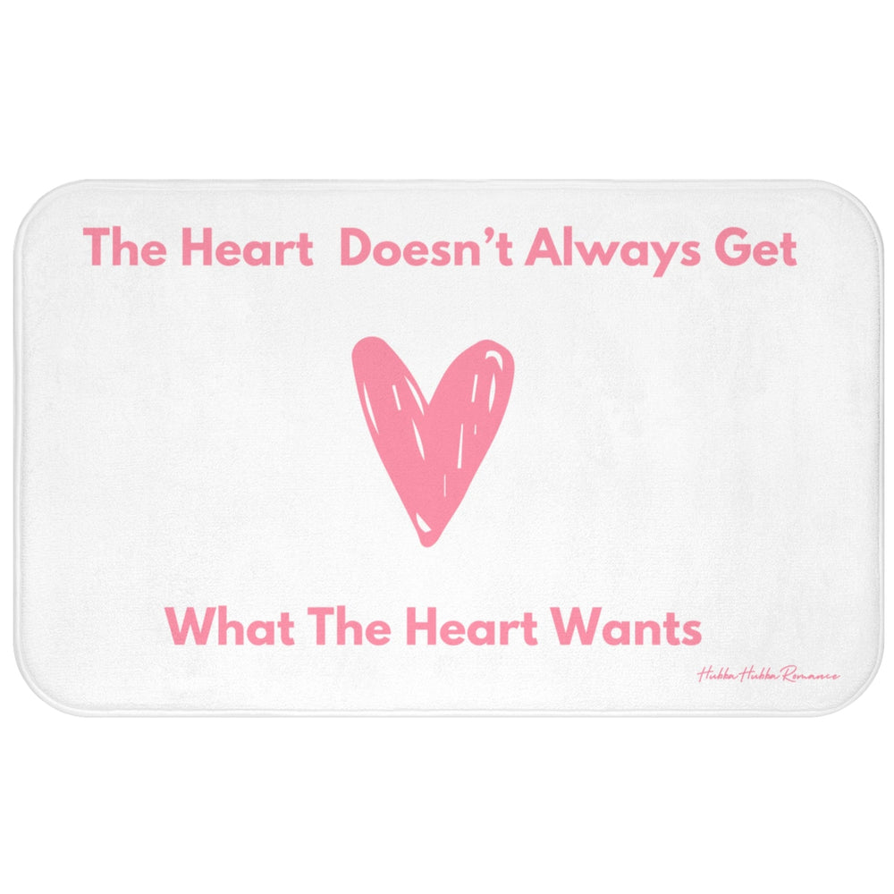 This bath mat features a clean white background with soft pink text and a large pink heart illustration at its center. The top line reads: “The Heart Doesn’t Always Get,” and the bottom line finishes the sentence: “What The Heart Wants.” The heart is hand-drawn in a casual, playful style. The overall design is minimalist and emotionally expressive. The mat features a soft, absorbent surface ideal for bathroom use, especially for romance readers who appreciate a touch of heartbreak with their hygiene. 