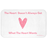 This bath mat features a clean white background with soft pink text and a large pink heart illustration at its center. The top line reads: “The Heart Doesn’t Always Get,” and the bottom line finishes the sentence: “What The Heart Wants.” The heart is hand-drawn in a casual, playful style. The overall design is minimalist and emotionally expressive. The mat features a soft, absorbent surface ideal for bathroom use, especially for romance readers who appreciate a touch of heartbreak with their hygiene. 