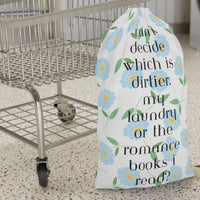 This design features light blue daisy-style flowers with yellow centers and green leaves on a white background. Black text reads: “Can’t decide which is dirtier: my laundry or the romance books I read?” for a cheeky, flirtatious message. The overall look is clean and pretty with a spicy twist for romance readers. There is a draw string enclosure and a large canvas carry strap. The bag is the smaller size and is displayed in front of a cart for context.
