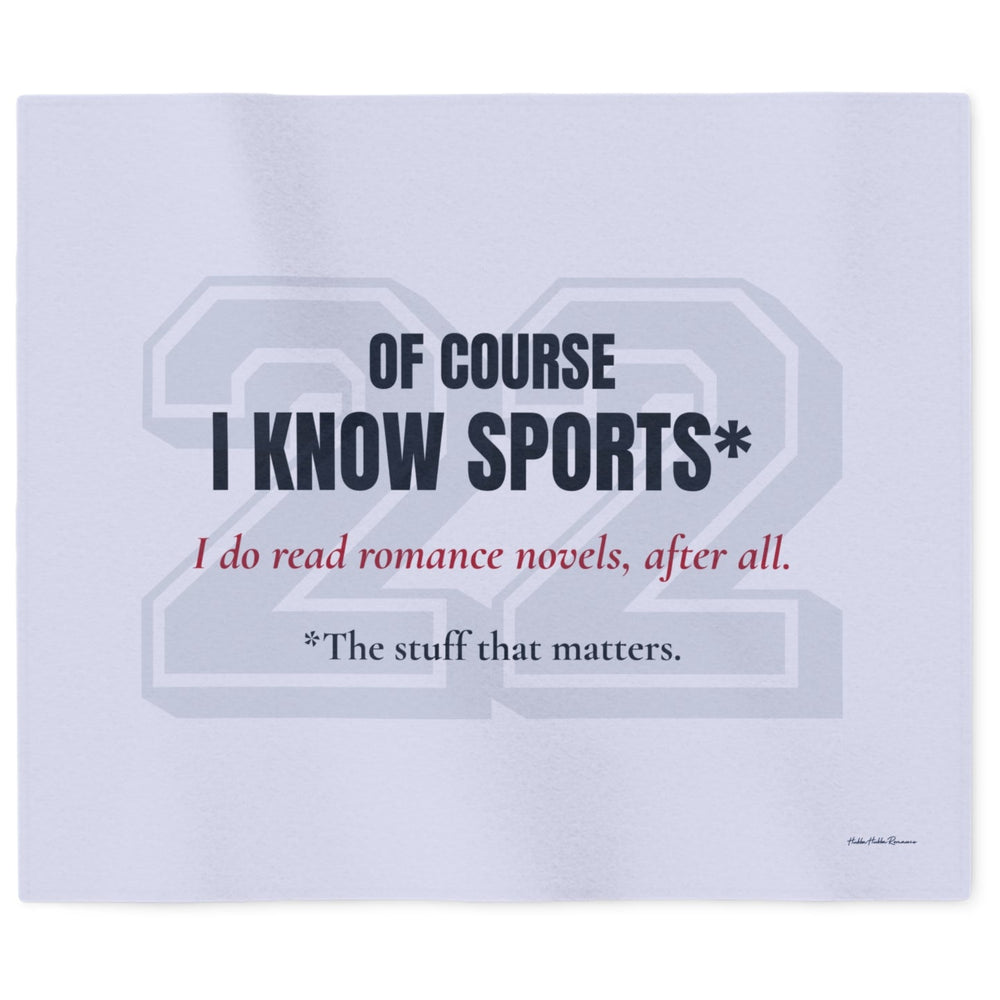 A light blueish gray sweatshirt blanket featuring a printed graphic with the text 'OF COURSE I KNOW SPORTS*' in dark navy, 'I do read romance novels, after all.' in red, and '*The stuff that matters in dark blue font. A large and very faded 22 in block numbers sits behind the text.