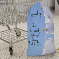 This laundry bag features two white tank tops pinned to a clothesline against a bright blue sky. To the left side of the t-shirts is a playful message in elegant black text: “Laundry. The second dirtiest thing that I do.” The visual combines clean imagery with cheeky humor, making it a fun and functional bag for readers who love steamy romance and tidy piles. The bag has a drawstring closure and a large canvas strap for carrying. This is the small version. It is displayed in front of a cart for size context