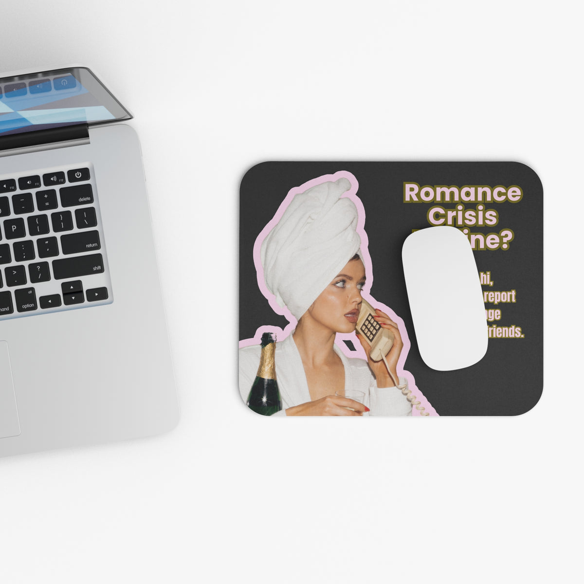 A rectangular mouse pad with rounded corners sits on a white surface next to a cropped silver laptop. The mat has a black background with a printed graphic of a woman wrapped in a white towel and holding a phone, plus the text 'Romance Crisis Line?' and a white computer mouse placed on top.