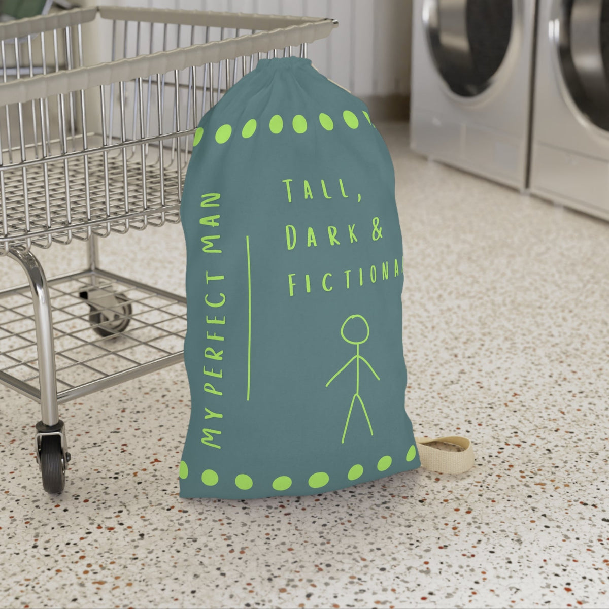 This laundry bag features a minimalist design on a muted greenish-gray background. A lime green stick figure stands next to bold lime green text that reads “Tall, Dark & Fictional,” with the words “My Perfect Man” printed vertically along the side. Rows of lime-green dots border the top and bottom of the bag. The bag includes a durable canvas carrying strap and a drawstring enclosure for secure, easy transport. This is the small version. It is in front of a cart for sizing context.