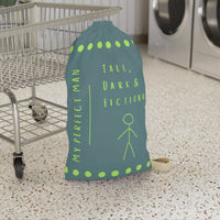 This laundry bag features a minimalist design on a muted greenish-gray background. A lime green stick figure stands next to bold lime green text that reads “Tall, Dark & Fictional,” with the words “My Perfect Man” printed vertically along the side. Rows of lime-green dots border the top and bottom of the bag. The bag includes a durable canvas carrying strap and a drawstring enclosure for secure, easy transport. This is the small version. It is in front of a cart for sizing context.