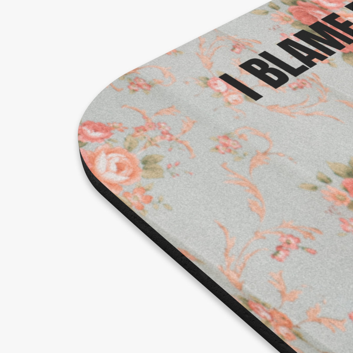 A close-up of a mouse pad, featuring a light gray background with a vintage floral pattern in pink, peach, and muted green. The top edge has the text 'I BLAME' printed in bold black letters, and the bottom edge has a black border.