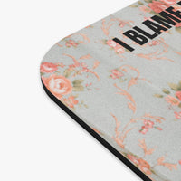A close-up of a mouse pad, featuring a light gray background with a vintage floral pattern in pink, peach, and muted green. The top edge has the text 'I BLAME' printed in bold black letters, and the bottom edge has a black border.