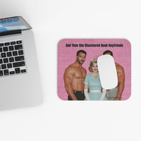 A rectangular printed mouse pad with rounded corners sits on a white desk beside a cropped silver laptop. The mouse pad features a pink textured background with a retro-style illustration of two shirtless men and a woman, plus the text 'And Then She Discovered Book Boyfriends.