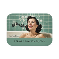 This bath mat features a retro-style image of a glamorous woman with styled dark hair, red lipstick, and a joyful smile, lounging in a bubble bath. Behind her is a sea-green tiled bathroom wall with soap bubbles floating around. Playful black text reads “Rub A Dub Dub.” At the bottom of the mat, centered over a seafoam green background strip, bold black text reads: “I Need A Man For My Tub.” The overall vibe is vintage pin-up with playful humor. This is the smaller version.
