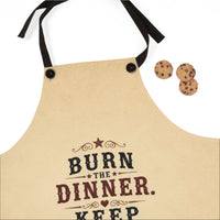 A beige kitchen apron with black neck and waist straps, featuring a western-style printed design that reads 'BURN THE DINNER. KEEP' in dark red and black lettering.