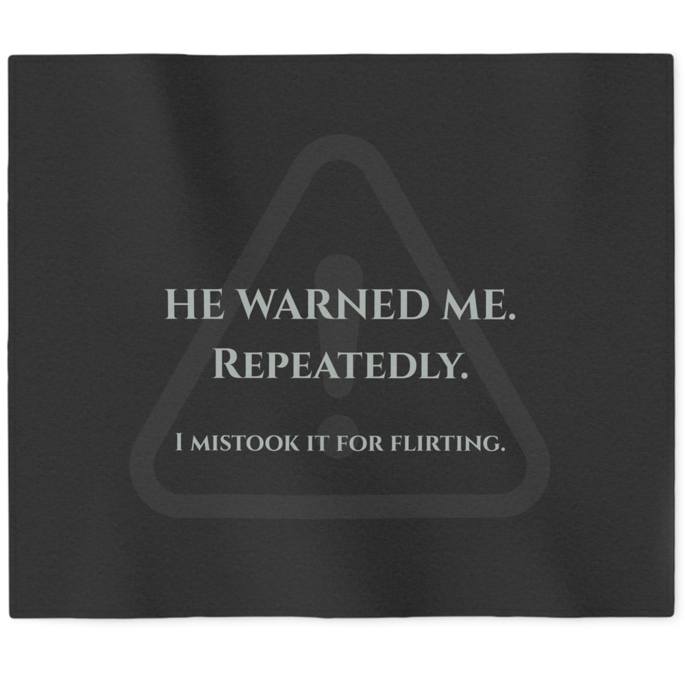 A black rectangular sweatshirt blanket with a gray warning-triangle graphic and the text 'HE WARNED ME. REPEATEDLY. I mistook it for flirting.