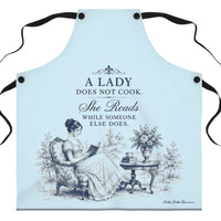 A light blue kitchen apron with black neck and waist ties, featuring a vintage illustration of a lady reading and the text 'A LADY DOES NOT COOK. She Reads WHILE SOMEONE ELSE DOES.