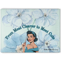 Glass cutting board featuring a retro cartoon-style woman in a blue off-shoulder dress and matching headband, holding a kitchen knife with a cheerful expression. She is surrounded by large watercolor-style blue flowers. Above her is the curved text: “From Meat Cleaver to Meet Cute.” Durable tempered glass with a textured surface and non-slip rubber feet. This is the larger size.