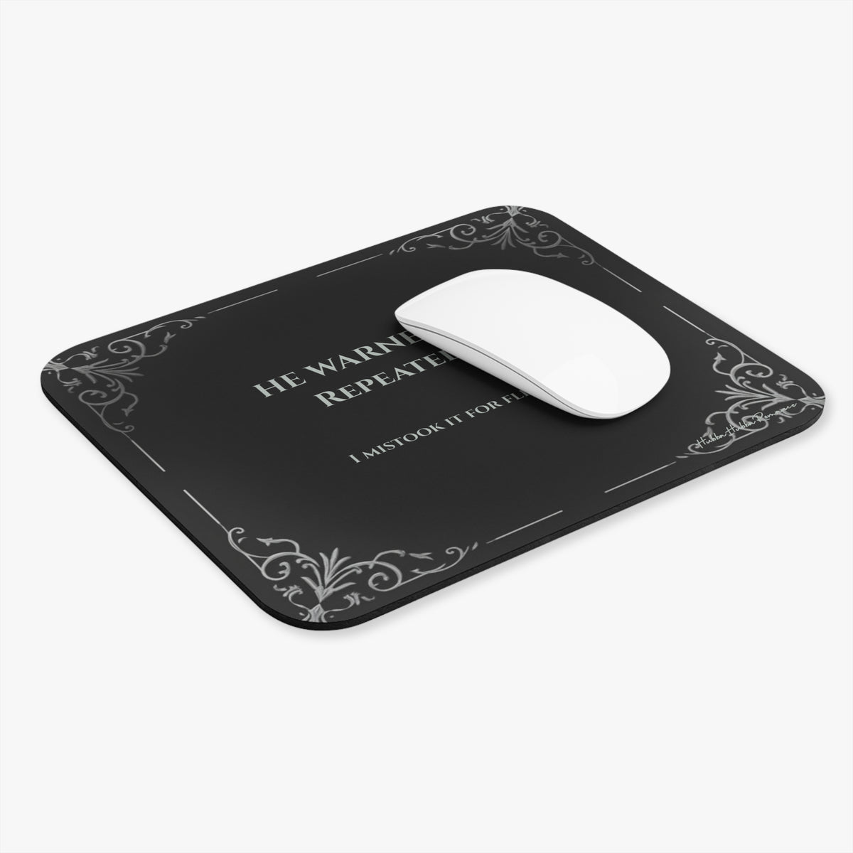 A white computer mouse sits on a rectangular black mouse pad with rounded corners, featuring light grey text and light gray filigree corner accents. The text  reads 'HE WARNED ME. REPEATEDLY. I mistook it for flirting.