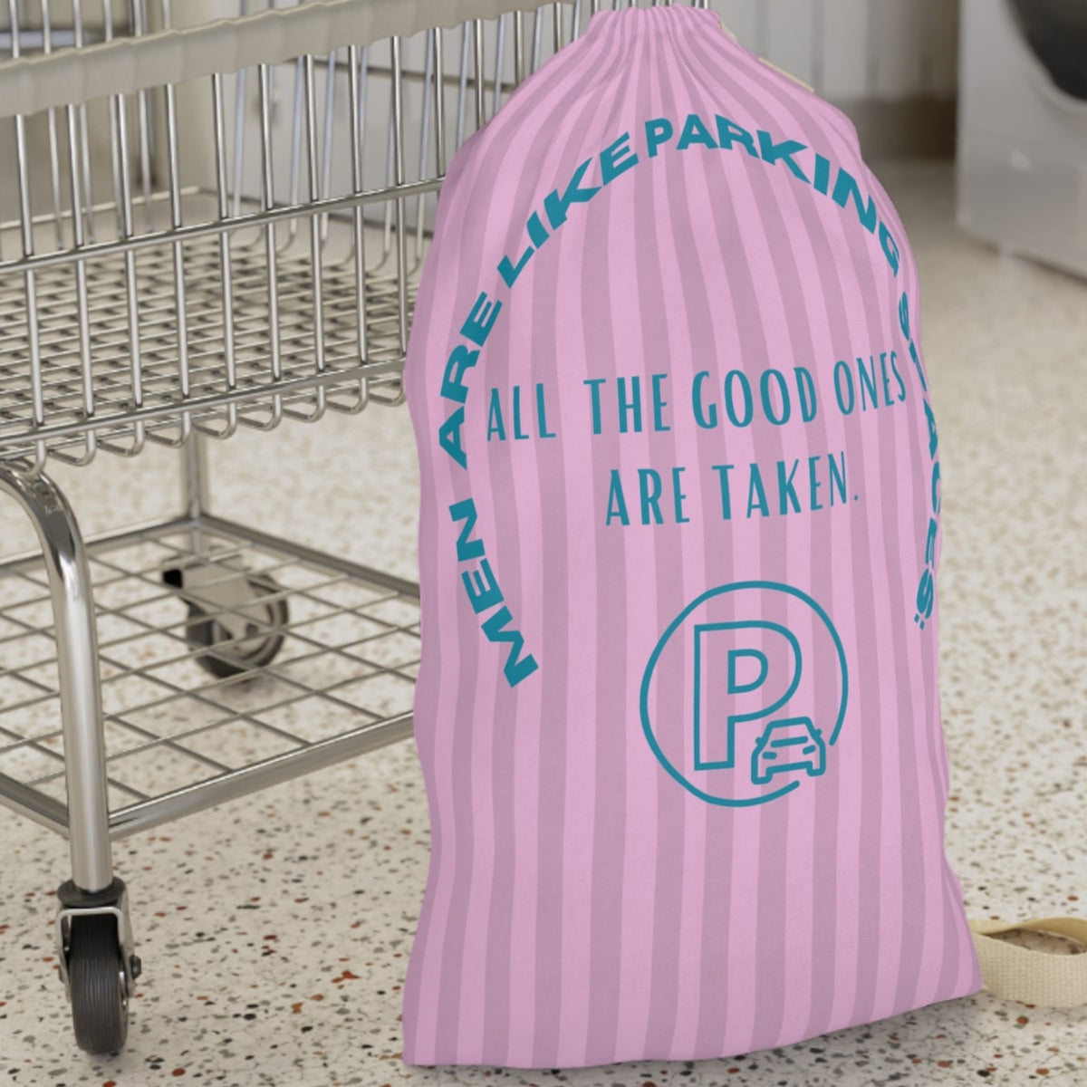 This laundry bag features a pink background with vertical pastel-pink stripes. Centered, bold, teal text reads: "All the good ones are taken." Above the text, in a large curved arc, it says: “Men are like parking spaces!” Below the quote is a parking symbol: a capital letter “P” inside a circle with a car icon in the corner. The bag includes a drawstring closure and a canvas carrying strap. The tone is playful, retro, and full of tongue-in-cheek charm. It is placed in front of a cart for size context