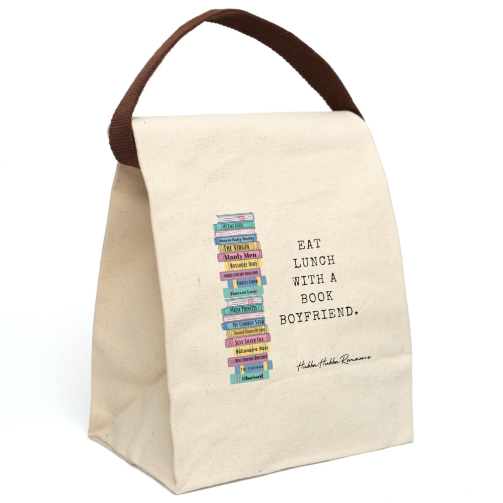 This canvas lunch bag features a playful stack of illustrated romance novels on the left side. Each book spine is brightly colored  with romance tropes as titles like “My Fake Fiancé,” “Secret Baby Daddy,” “Mafia Princess,” “Sexy Silver Fox,” and more. To the right of the stack, bold black typewriter-style text reads: “EAT LUNCH WITH A BOOK BOYFRIEND.” The background is a natural canvas. There is a brown canvas strap for carrying, which is displayed upright.