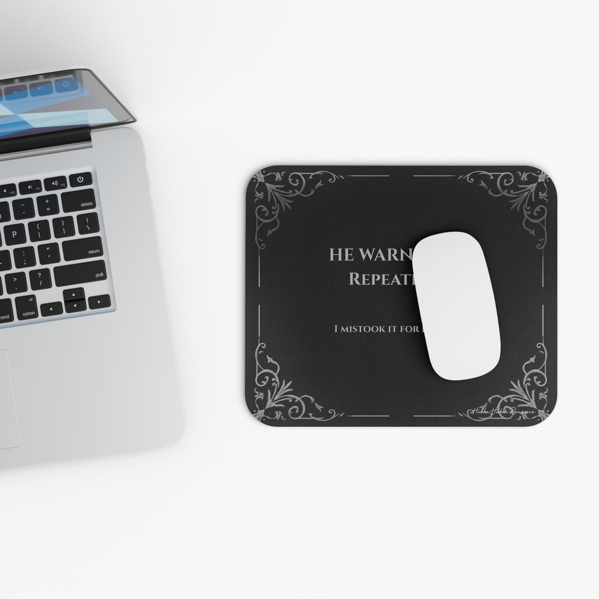 A white computer mouse sits on a rectangular black mouse pad with rounded corners, featuring light grey text and light gray filigree corner accents. The text  reads 'HE WARNED ME. REPEATEDLY. I mistook it for flirting. The mouse pad is located next to a laptop.