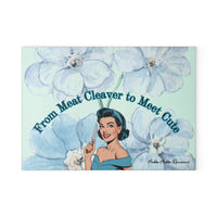 Glass cutting board featuring a retro cartoon-style woman in a blue off-shoulder dress and matching headband, holding a kitchen knife with a cheerful expression. She is surrounded by large watercolor-style blue flowers. Above her is the curved text: “From Meat Cleaver to Meet Cute.” Durable tempered glass with a textured surface and non-slip rubber feet. This is the smaller size.