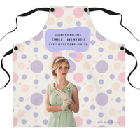 A beige apron with pastel pink and purple polka dots and black straps stands in a modern kitchen. The apron features a vintage-style illustration of a glamorous woman in a mint green dress, pearl necklace, and gloves. Above her, a purple speech bubble contains the text: "I like my recipes simple… and my book boyfriends complicated."
