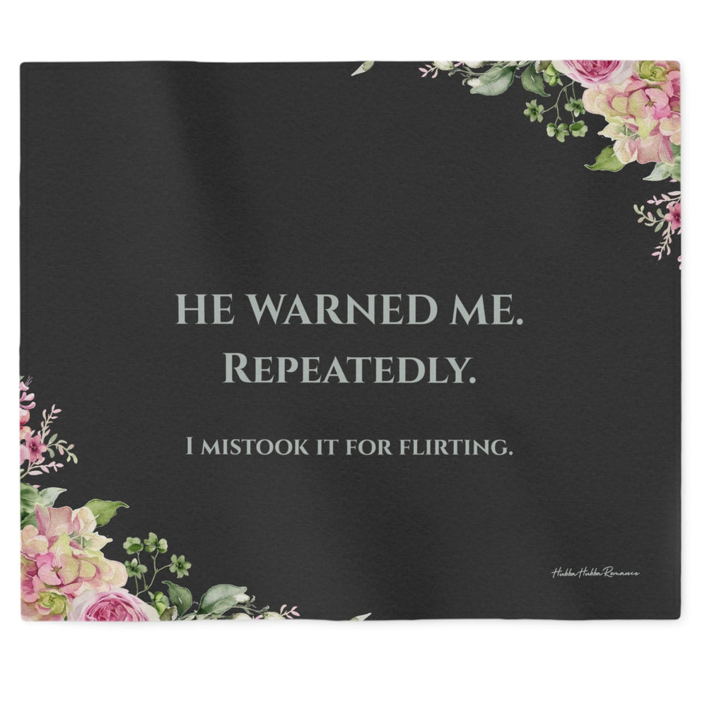  a dark charcoal sweatshirt blanket with light grey text that reads 'HE WARNED ME. REPEATEDLY. Then in smaller font, "I Mistook It For Flirting". There is a small floral motif in the lower left and upper right corner.