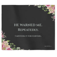  a dark charcoal sweatshirt blanket with light grey text that reads 'HE WARNED ME. REPEATEDLY. Then in smaller font, "I Mistook It For Flirting". There is a small floral motif in the lower left and upper right corner.