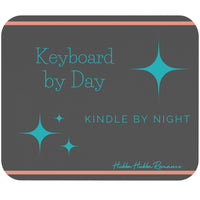 Mouse pad with a gray background and a thin orange stripe at the top and bottom of the mouse pad. Scattered across the pad are retro star shapes in a turquoise color. Turquoise text reads: “Keyboard by day” in the upper left quadrant and “Kindle by Night” in the lower right quadrant.
