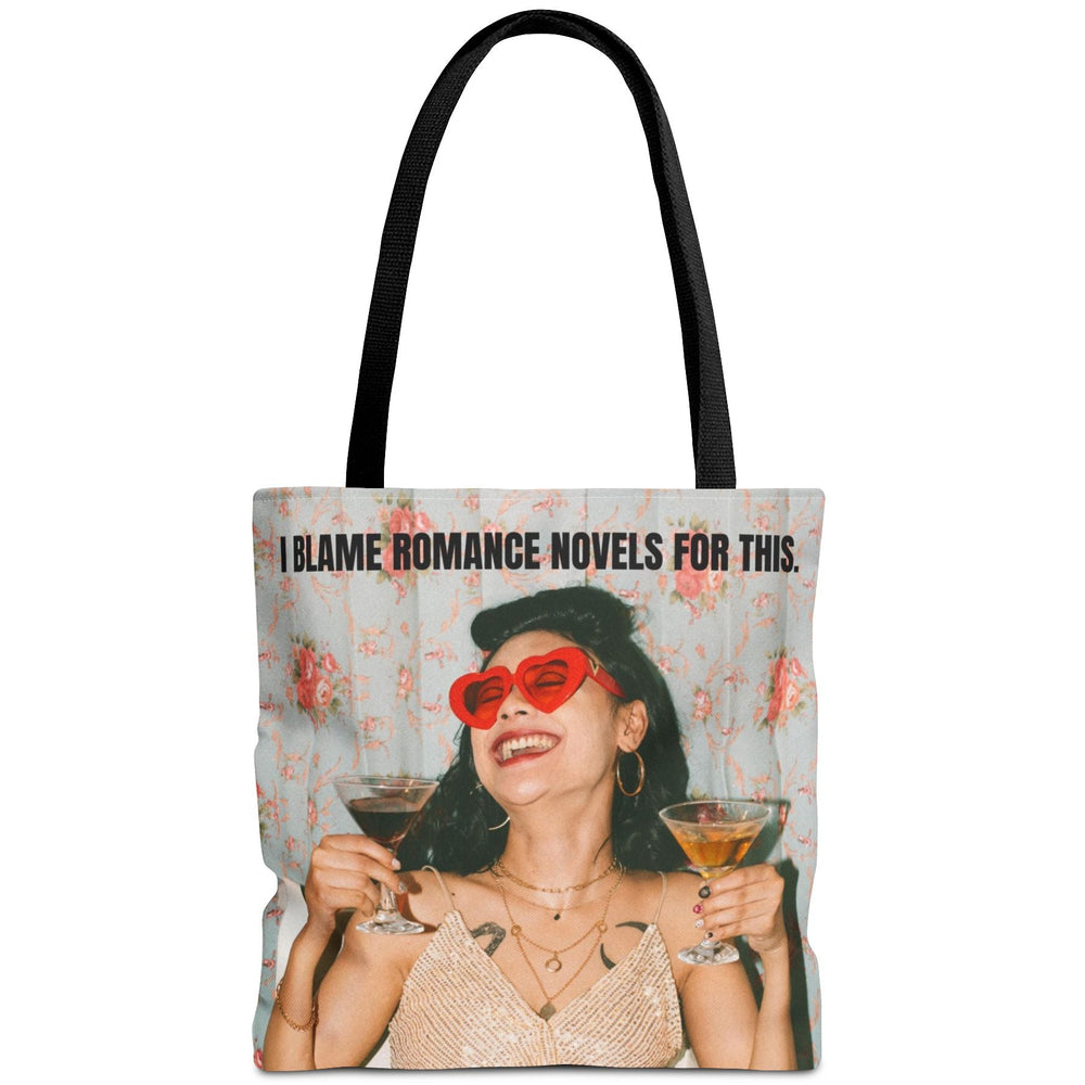 A tote bag with black handles featuring a printed graphic of a woman in a beige dress, red heart-shaped sunglasses, and a black hat, holding two cocktails. The text above reads 'I BLAME ROMANCE NOVELS FOR THIS.