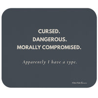 A rectangular dark slate-gray mouse pad with rounded corners, featuring the text 'CURSED. DANGEROUS. Morally Compromised. Apparently I have a type in cream letters.
