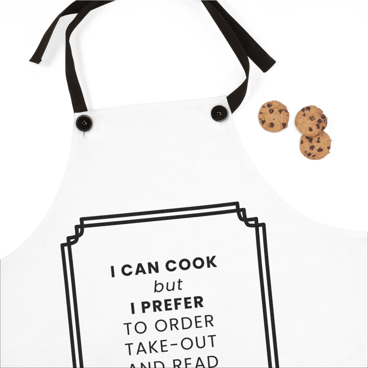 A white kitchen apron with black neck and waist straps, featuring a black graphic frame and the text 'I CAN COOK but I PREFER TO ORDER TAKE-OUT AND READ' on the front. Three chocolate chip cookies are placed to the right on a clean white background.