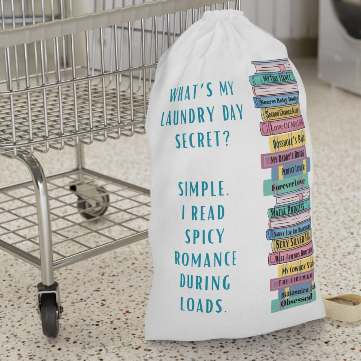 This is an image of an off white laundry bag with a drawstring enclosure at the top and a large canvas carrying strap on the side. The bag has an image of a large stack of romance books with various spicy titles. Next to the stack is text that says: What's My Laundry Secret? Simple. I read Spicy Romance During Loads. This is the smaller bag and is displayed in front of a laundry cart for size context.
