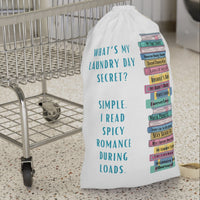 This is an image of an off white laundry bag with a drawstring enclosure at the top and a large canvas carrying strap on the side. The bag has an image of a large stack of romance books with various spicy titles. Next to the stack is text that says: What's My Laundry Secret? Simple. I read Spicy Romance During Loads. This is the smaller bag and is displayed in front of a laundry cart for size context.
