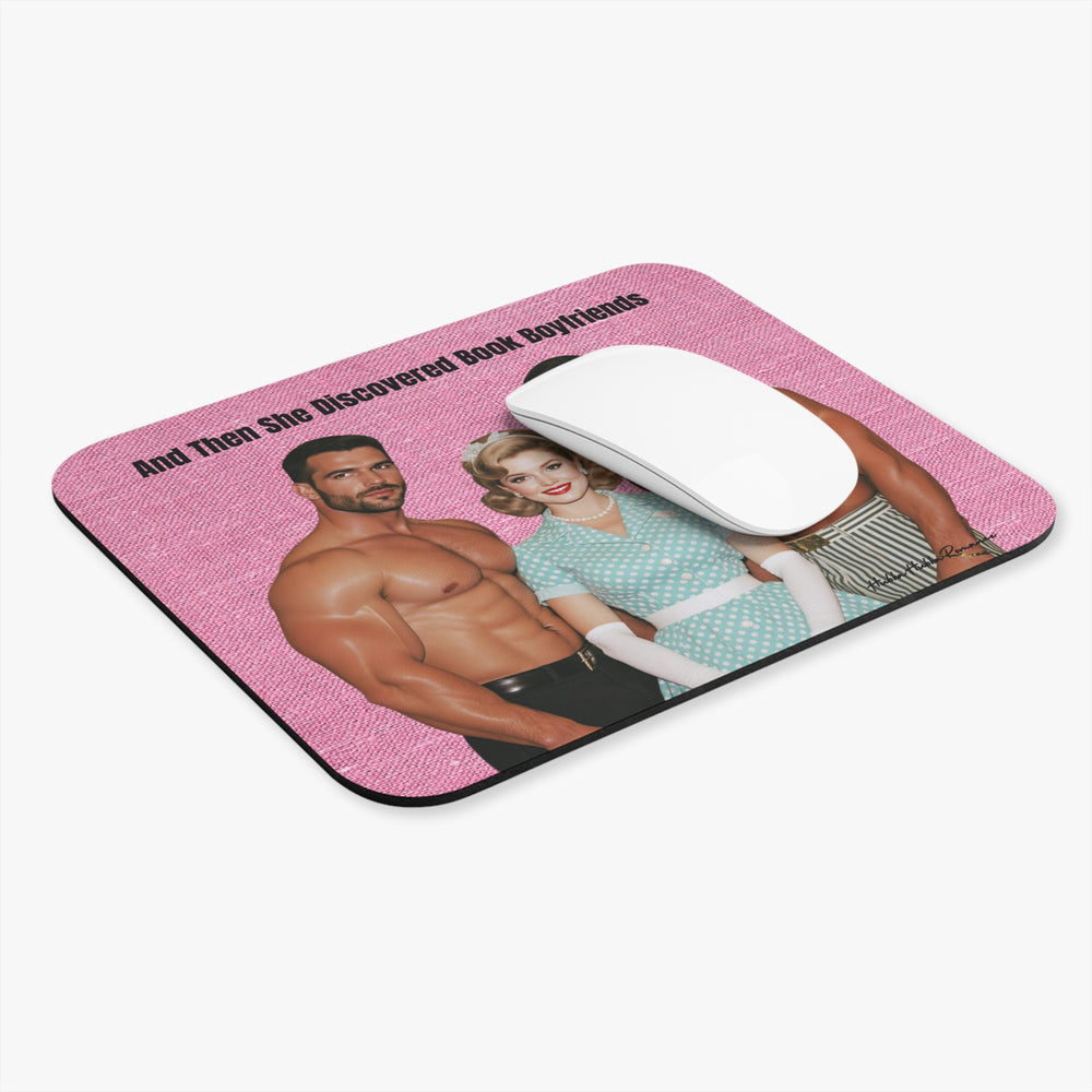A rectangular computer mouse pad with rounded corners, featuring a pink background and a retro-style printed illustration of a shirtless man and a woman in a turquoise dress. A white computer mouse is placed on top of the pad. The text reads "And then she discovered book boyfriends."