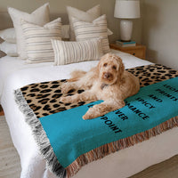 The blanket is displayed neatly folded on a bed with a white bedspread and striped pillows and a large white dog laying on it.
