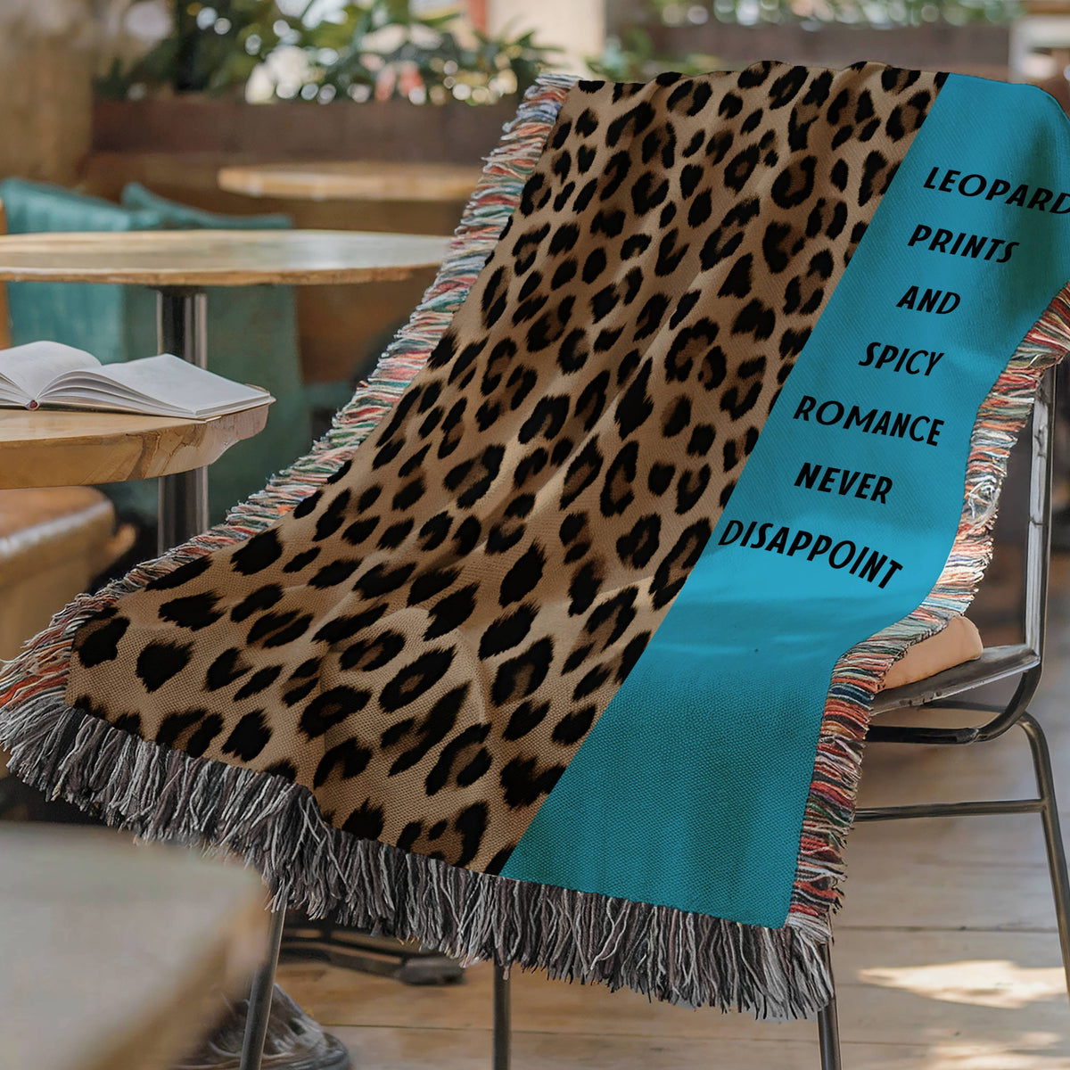 The blanket is displayed lying on a chair in a coffee shop setting with an open book and a cup of coffee on a small round table.
