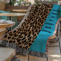 The blanket is displayed lying on a chair in a coffee shop setting with an open book and a cup of coffee on a small round table.
