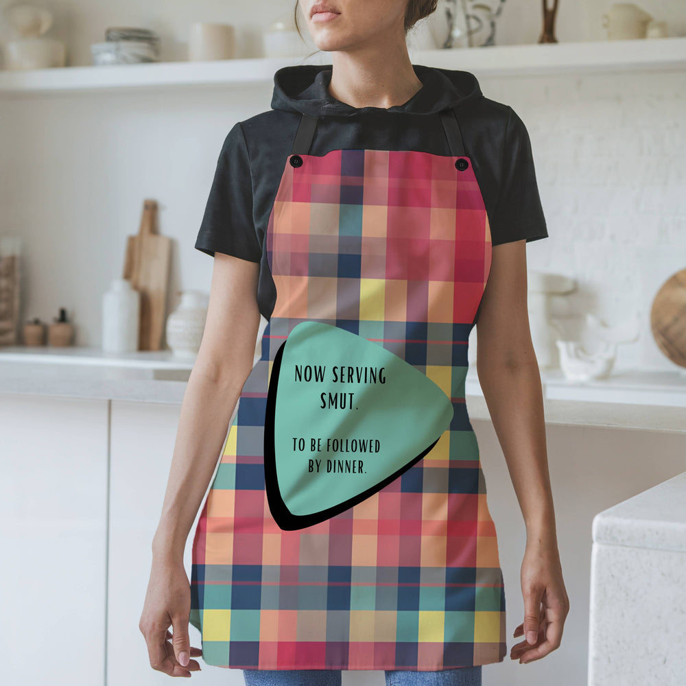 Pix text: A woman wearing a colorful plaid apron with black straps stands in a modern kitchen. The apron features a retro-inspired teal and black geometric shape containing the text: "Now serving smut. To be followed by dinner." The background includes white shelves, cutting boards, and kitchen items.