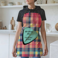 Pix text: A woman wearing a colorful plaid apron with black straps stands in a modern kitchen. The apron features a retro-inspired teal and black geometric shape containing the text: "Now serving smut. To be followed by dinner." The background includes white shelves, cutting boards, and kitchen items.