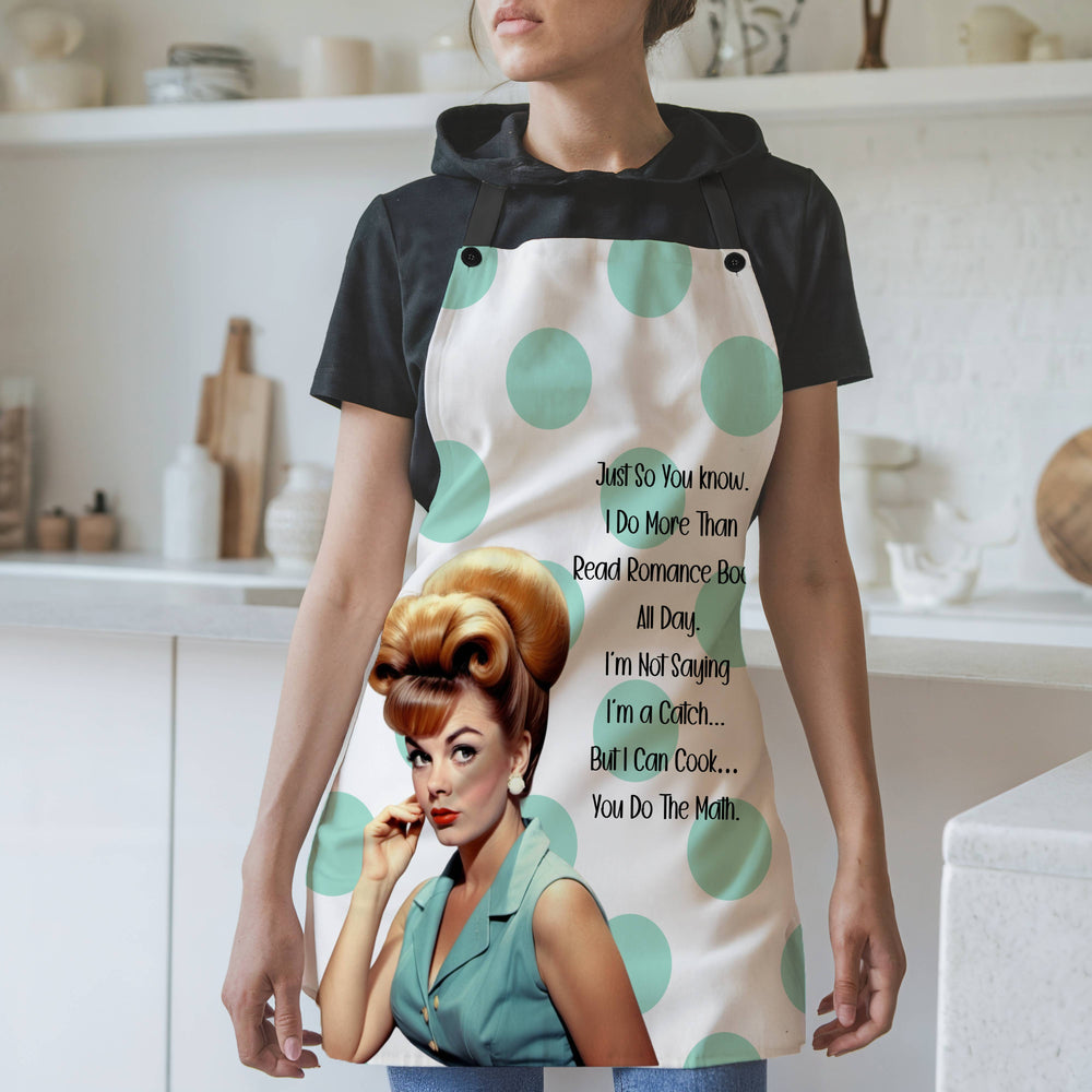 A woman wearing a white apron with large mint-green polka dots and black straps stands in a modern kitchen. The apron showcases a retro-style illustration of a glamorous 1960s woman with red hair styled in an updo, adorned in a teal dress and pearl earrings. The text reads: "Just so you know, I do more than read romance novels all day. I'm not saying I'm a catch... but I can cook... you do the math." The background features white shelves, cutting boards, and various kitchen items. 