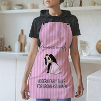 A woman wearing a pink striped apron with black straps stands in a modern kitchen. The apron features the phrase "Romance novels:" in elegant script, followed by "Modern fairy tales for grown-ass women" in bold lettering. Below the text, there is an illustration of a bride and groom in a dramatic dip, with the groom kissing the bride. The background includes white shelves, cutting boards, and kitchen items.