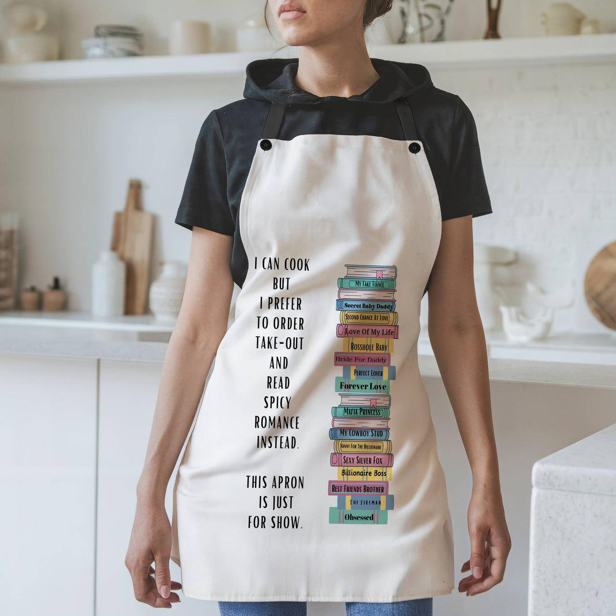 : A woman wearing a white apron with black straps stands in a modern kitchen. The apron features a vertical stack of illustrated romance novels with colorful spines. To the left of the books, black text reads: "I can cook, but I prefer to order take-out and read spicy romance instead. This apron is just for show." The background includes white shelves, cutting boards, and kitchen items.
