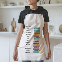 : A woman wearing a white apron with black straps stands in a modern kitchen. The apron features a vertical stack of illustrated romance novels with colorful spines. To the left of the books, black text reads: "I can cook, but I prefer to order take-out and read spicy romance instead. This apron is just for show." The background includes white shelves, cutting boards, and kitchen items.