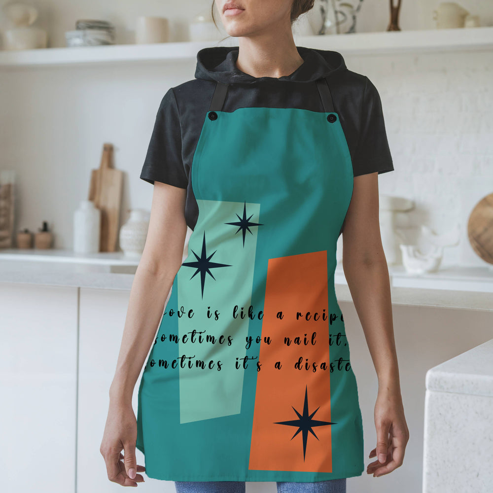A woman wearing a teal apron with black straps stands in a modern kitchen. The apron features a retro mid-century modern design with abstract teal and orange shapes, accented by black starbursts. The text across the apron reads: "Love is like a recipe. Sometimes you nail it, sometimes it’s a disaster." The background includes white shelves, cutting boards, and kitchen items.
