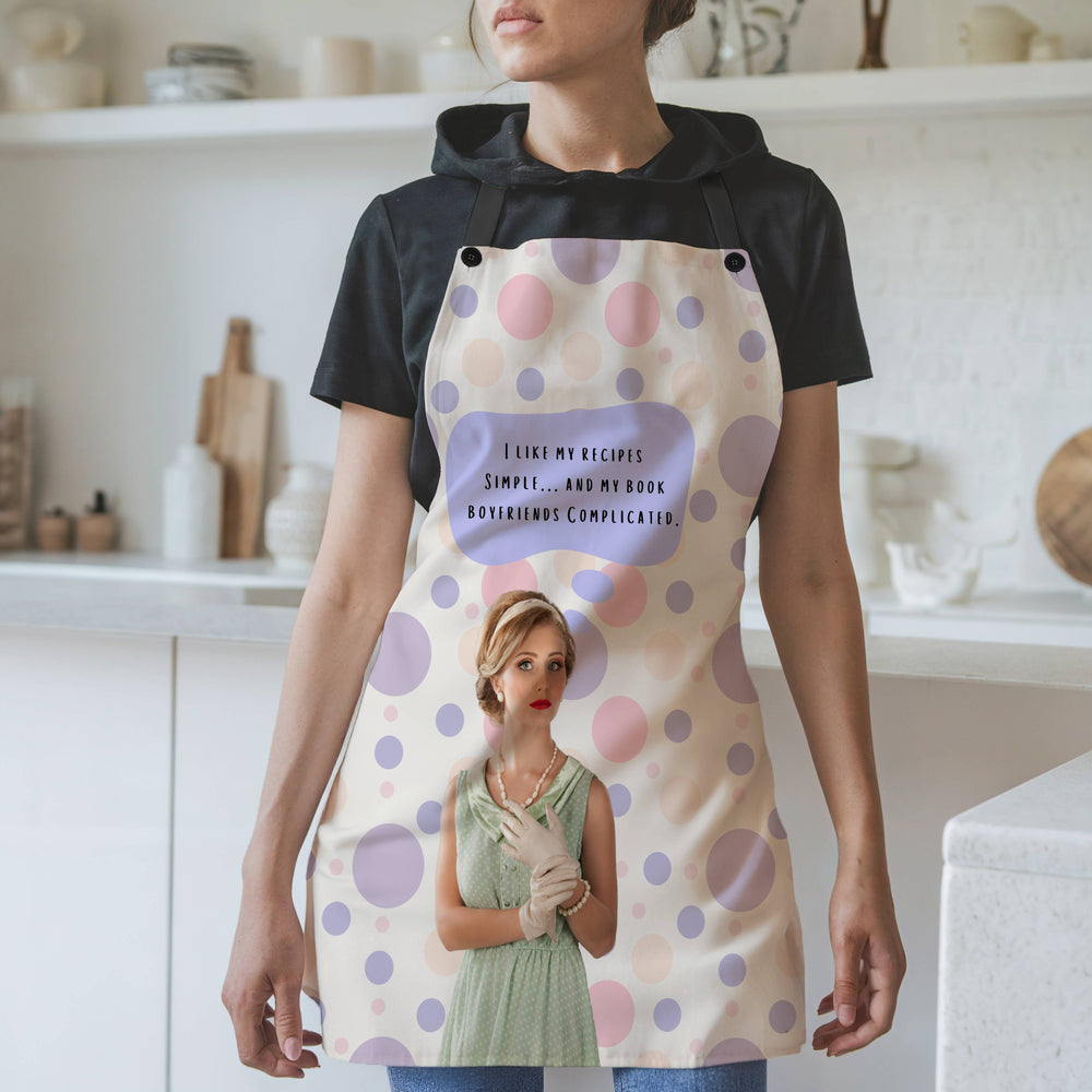 A woman wearing a beige apron with pastel pink and purple polka dots and black straps stands in a modern kitchen. The apron features a vintage-style illustration of a glamorous woman in a mint green dress, pearl necklace, and gloves. Above her, a purple speech bubble contains the text: "I like my recipes simple… and my book boyfriends complicated." The background includes white shelves, cutting boards, and kitchen items.