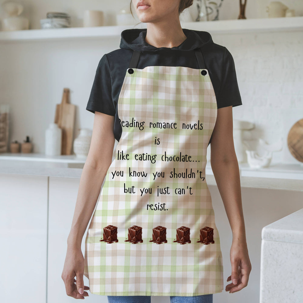 A woman wearing an apron with a white background and a pink and light green apron with black straps stands in a modern kitchen. The apron features black text that reads: "Reading romance novels is like eating chocolate… you know you shouldn’t, but you just can’t resist." Below the text, there is a row of illustrated chocolate pieces melting at the bottom. The background includes white shelves, cutting boards, and kitchen items.
