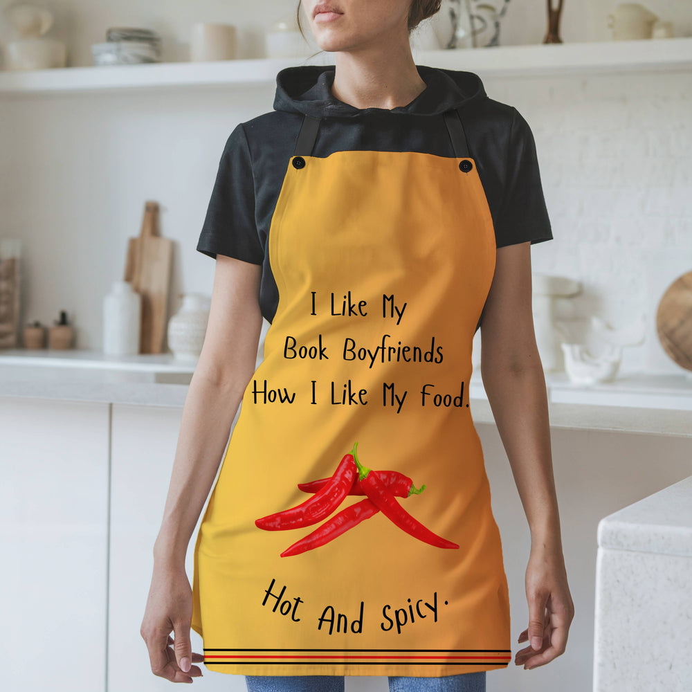 A woman wearing an orangy yellow apron with black straps stands in a modern kitchen. The apron features the phrase, "I like my book boyfriends how I like my food. Hot and spicy." in black text. Below the text, there is an illustration of three red chili peppers. The bottom of the apron has a decorative striped trim. The background includes white shelves, cutting boards, and kitchen items.