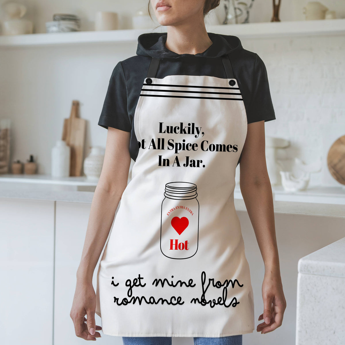 A woman wearing a white apron with black straps and a humorous design stands in a modern kitchen. The apron features the phrase, "Luckily, not all spice comes in a jar," with an illustration of a spice jar labeled "Hot" and "Extra Heat Level" inside a red heart. Below, the text reads, "I get mine from romance novels" in a playful script. The background includes white shelves, cutting boards, and kitchen items.