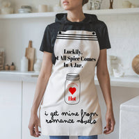 A woman wearing a white apron with black straps and a humorous design stands in a modern kitchen. The apron features the phrase, "Luckily, not all spice comes in a jar," with an illustration of a spice jar labeled "Hot" and "Extra Heat Level" inside a red heart. Below, the text reads, "I get mine from romance novels" in a playful script. The background includes white shelves, cutting boards, and kitchen items.