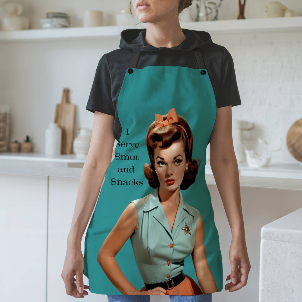 A woman wearing a teal-colored apron with black straps stands in a modern kitchen. The apron features a vintage-style illustration of a glamorous 1950s woman with red hair styled in curls. She wears a mint green blouse with a sheriff’s star brooch, an orange skirt, and a matching bow in her hair. The text beside her reads: "I Serve Smut and Snacks." The background includes white shelves, cutting boards, and kitchen items.