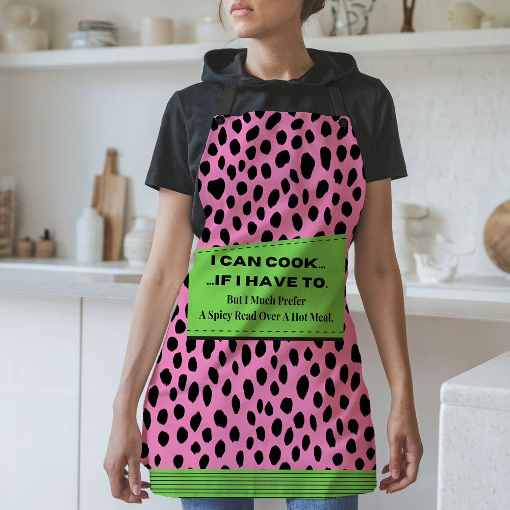 A woman wearing a bold pink and black leopard-print apron with black straps stands in a modern kitchen. The apron features a green patch with the text: "I CAN COOK... ...IF I HAVE TO. But I Much Prefer A Spicy Read Over A Hot Meal." The bottom of the apron has a coordinating green and black striped trim. The background includes white shelves, cutting boards, and kitchen items.