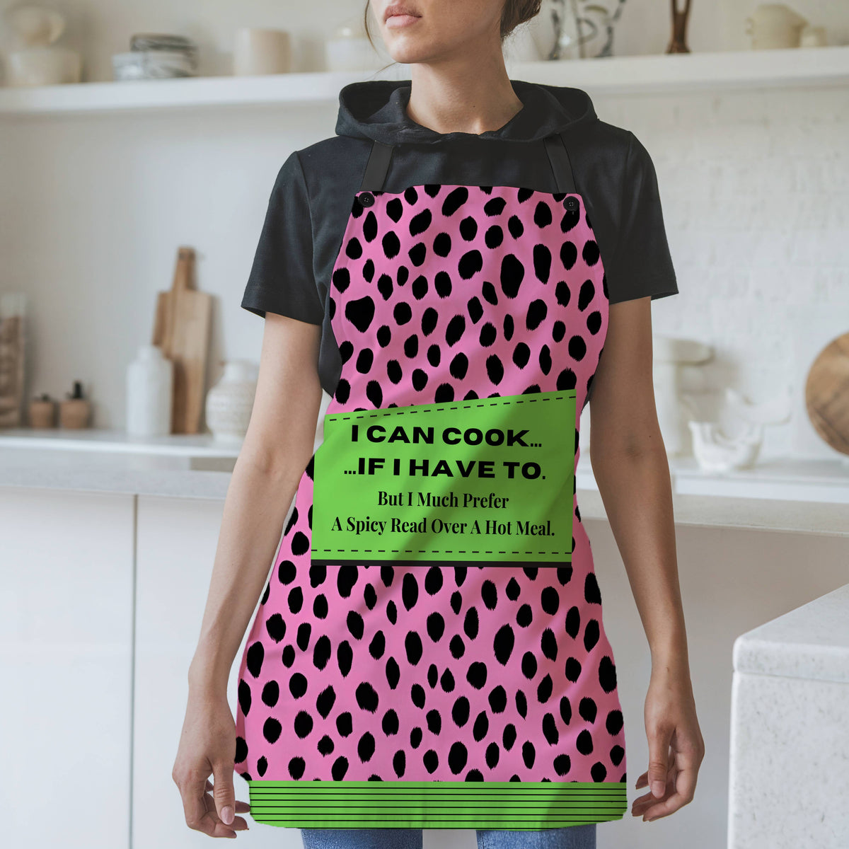 A woman wearing a bold pink and black leopard-print apron with black straps stands in a modern kitchen. The apron features a green patch with the text: "I CAN COOK... ...IF I HAVE TO. But I Much Prefer A Spicy Read Over A Hot Meal." The bottom of the apron has a coordinating green and black striped trim. The background includes white shelves, cutting boards, and kitchen items.