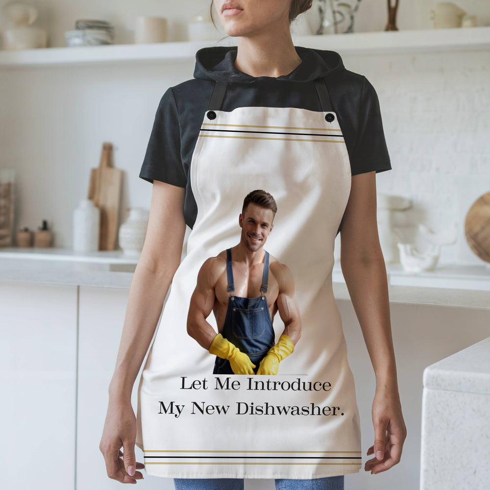 A woman wearing a white apron with black straps stands in a modern kitchen. The apron features an image of a muscular, shirtless man wearing denim overalls, yellow rubber gloves, and smiling. Below the image, the text reads: "Let Me Introduce My New Dishwasher." The apron has thin black and gold stripes along the top and bottom edges. The background includes white shelves, cutting boards, and kitchen items.